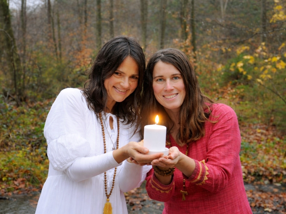 Yogita und Satyadevi halten gemeinsame eine weiße Kerze. Sie tragen weiße beziehungsweise rote Kleider. Sie lächeln in die Kamera.