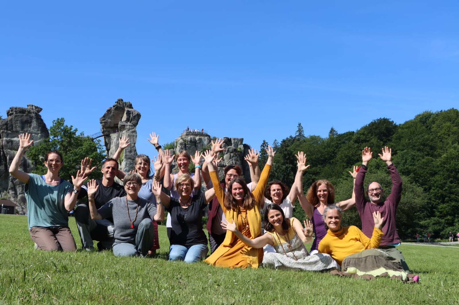 Gruppenfoto vor den Externsteinen. Mehrere Menschen sitzen auf dem Boden, lachen und winken in die Kamera.