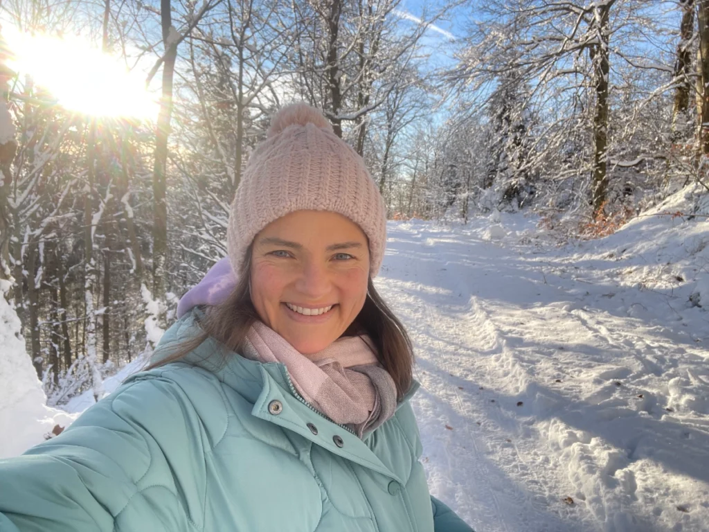 Selfie von Yogita in einem verschneiten Wald. Die Sonne strahlt durch die Bäume.