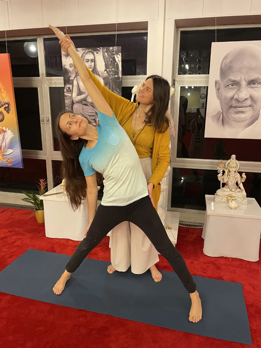 Yogita, die eine individuelle Yogastunde mit einer Yogaschülerin durchführt. Die Yogaschülerin steht barfuß auf einer Matte und neigt sich zur Seite. Yogita dehnt einen Arm der Schülerin zur Seite.