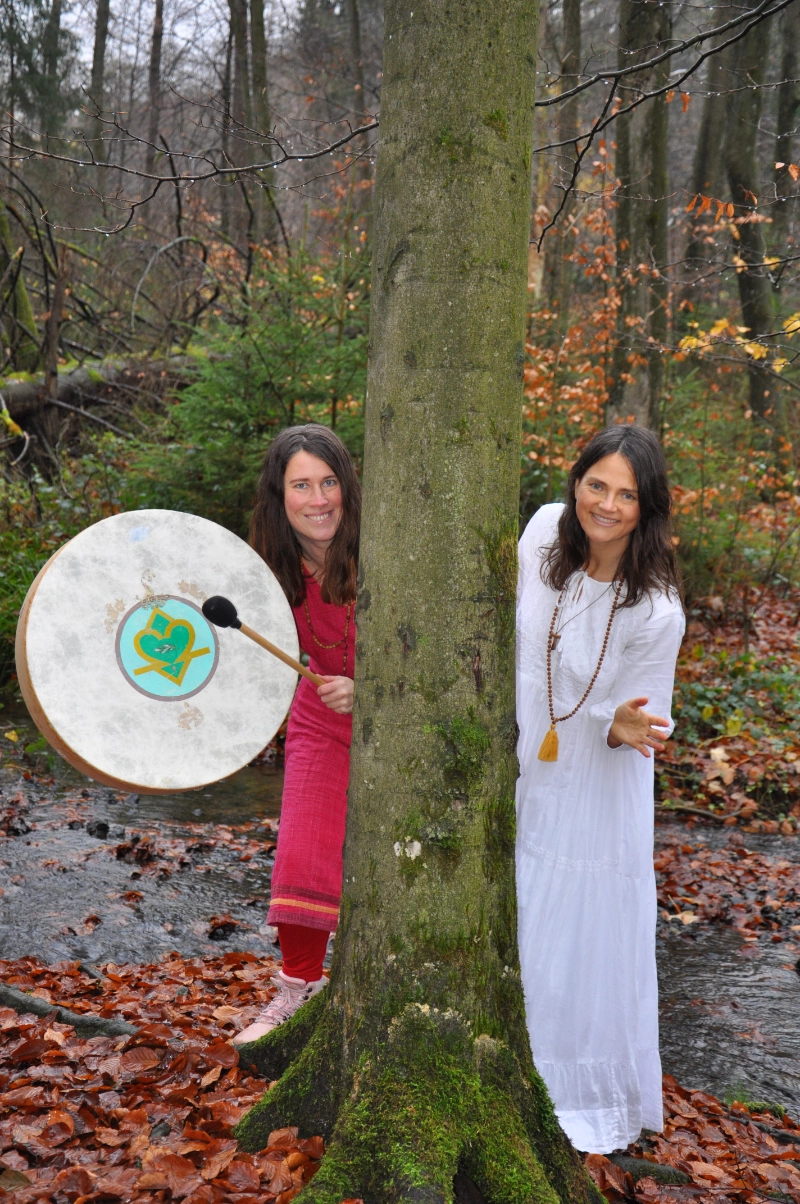 Satyadevi und Yogita schauen hinter einem Baum hervor. Beide tragen lange Kleider in Rot beziehungsweise Weiß. Satyadevi hält eine große Trommel mit Schlegel in den Händen.